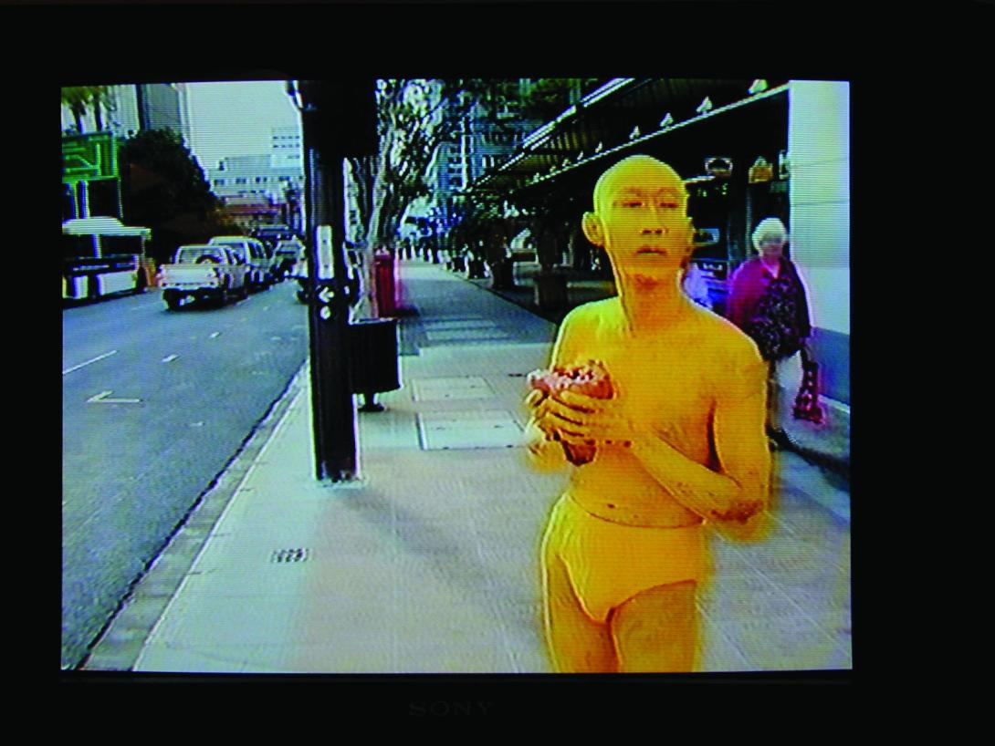 A photograph of a CRT television screen playing a video, in which a bald man painted bright yellow, wearing only yellow underpants, carries a red heart through the streets of Brisbane.