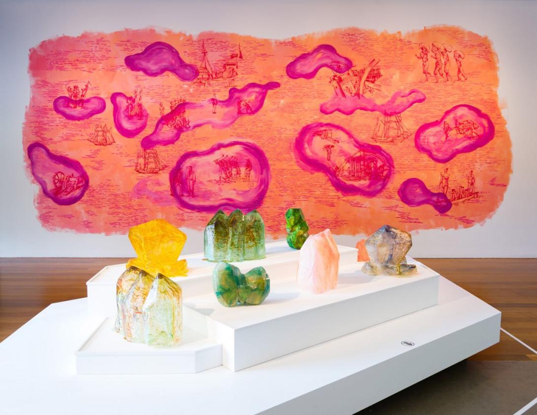 An installation view of sculptures that look like large, bright green, yellow and pink gems. Behind them, a bright orange and pink mural is painted directly on the wall.