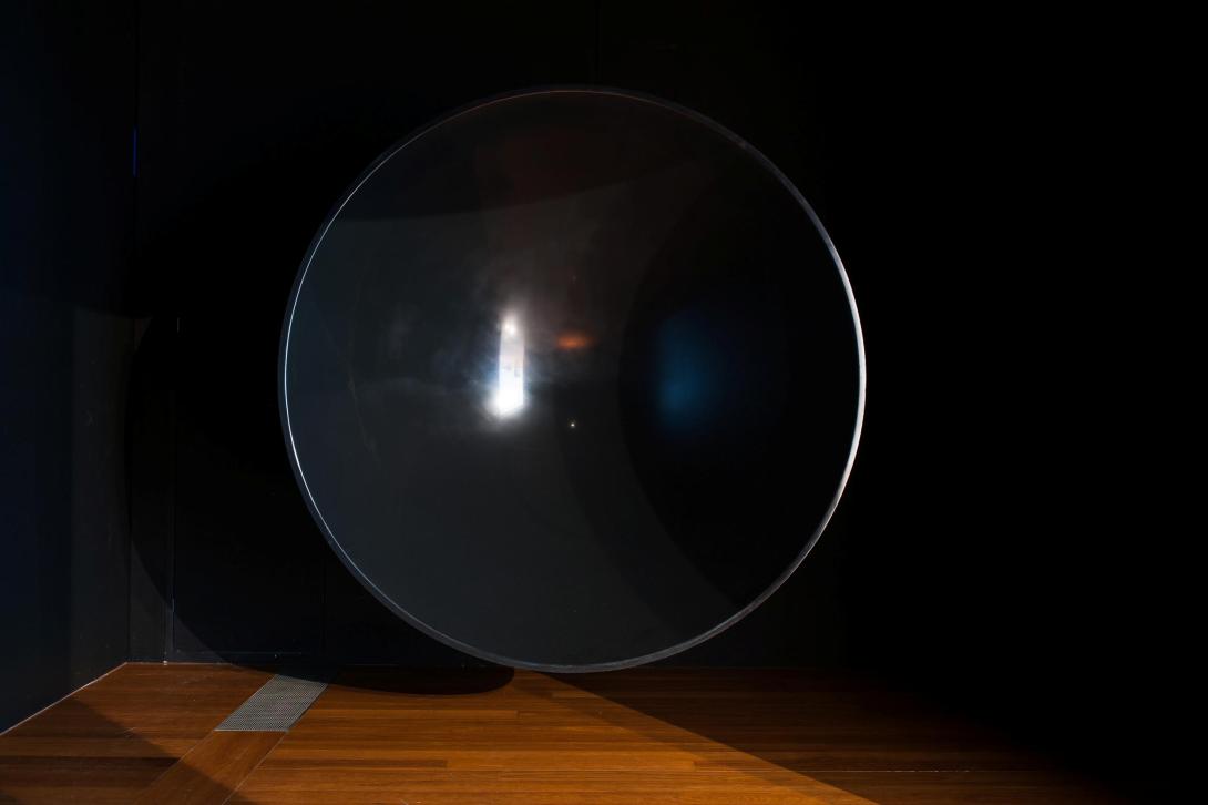 An installation view of a large concave, round black sculpture; the background is dark and the floor is polished hardwood.