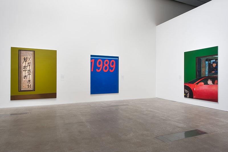 An installation view of three brightly coloured oil paintings hung on white walls in a gallery space.