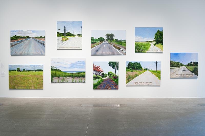 An installation view of nine works on a white gallery wall; each work depicts a road or landscape.