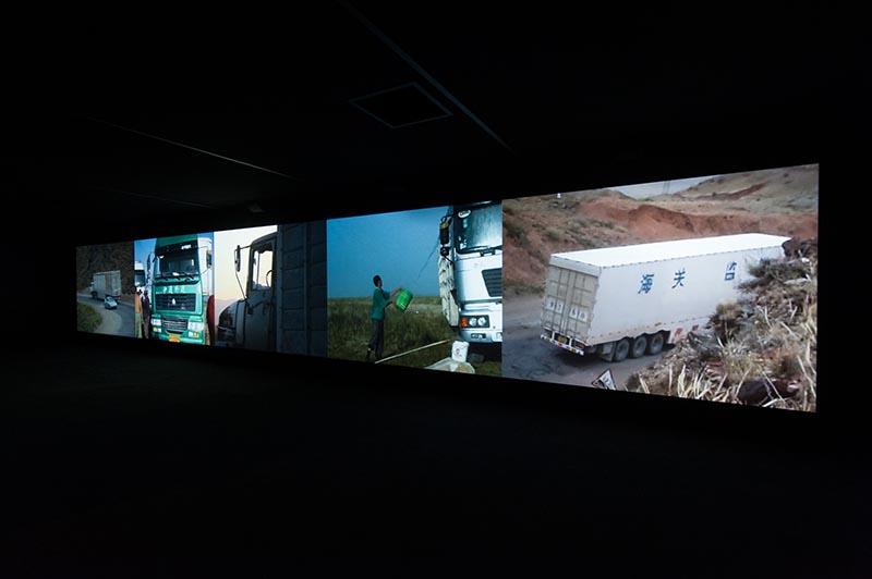 An installation view of a video work featuring trucks travelling through various landscapes.