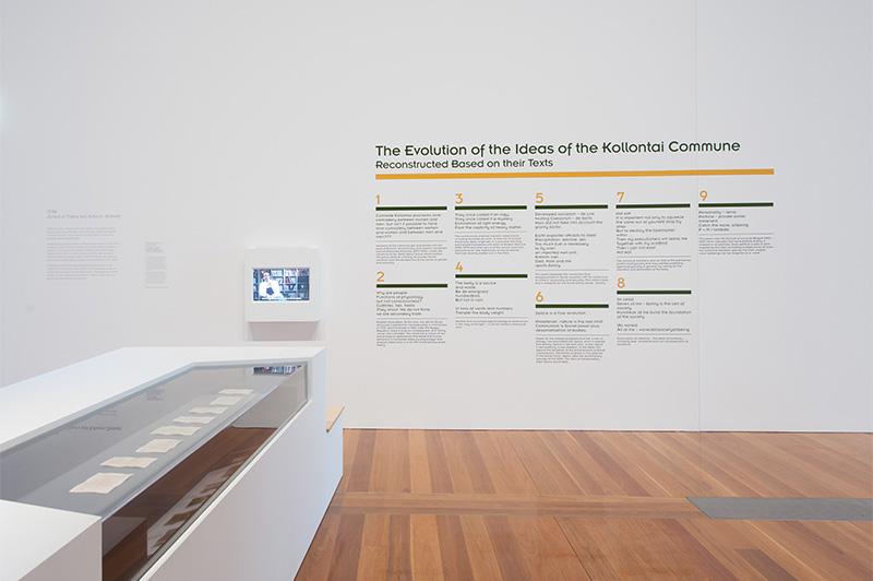 An installation view of a gallery space with works installed in a glass cabinet at left, with text applied to the wall in the background.