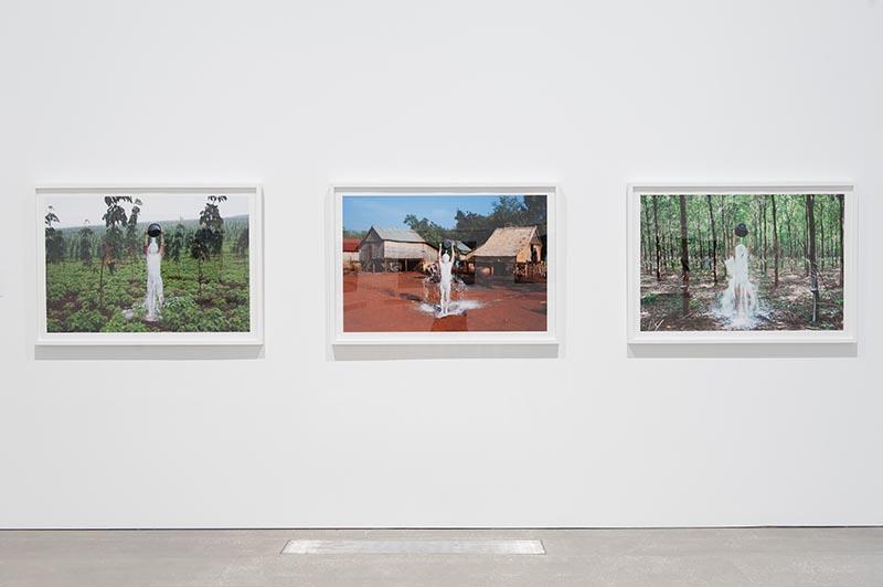 An installation view of three photographs on a white gallery wall; each features a man standing in a different environs tipping white rubber sap over his body.