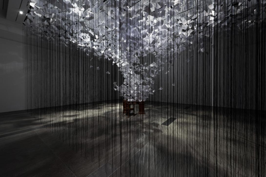 An installation view of a work that fills a whole room; at the centre, a desk and empty chair have thousands of papers appearing to fly off of them in a spiral. Suspended around this scene is a web of black threads.