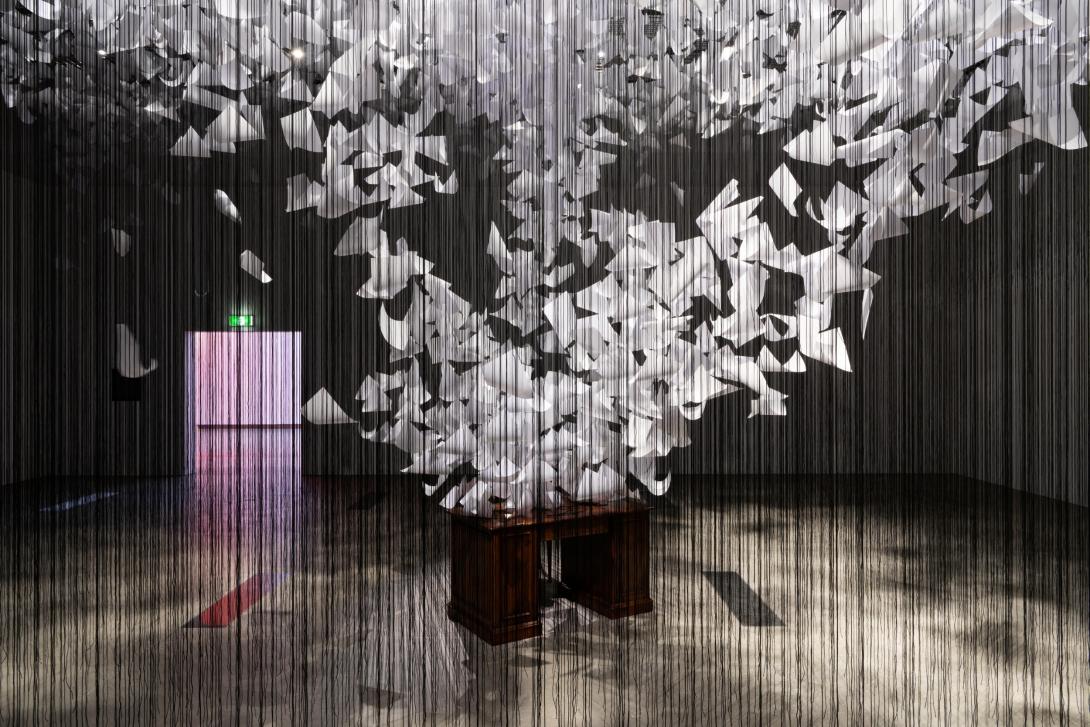 An detail view of a room-filling installation: seen through what appears as a curtain of black threads, a desk and empty chair have thousands of papers appearing to fly off of them in a tornado-like spiral. In the background, the exit from the gallery space is lit in pink.