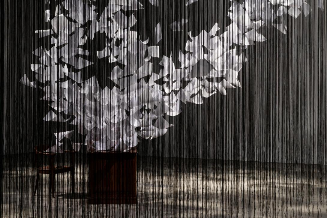 A detail view of a room-filling installation: seen through what appears as a curtain of black threads, a desk and empty chair have thousands of papers appearing to fly off of them in a tornado-like spiral. 