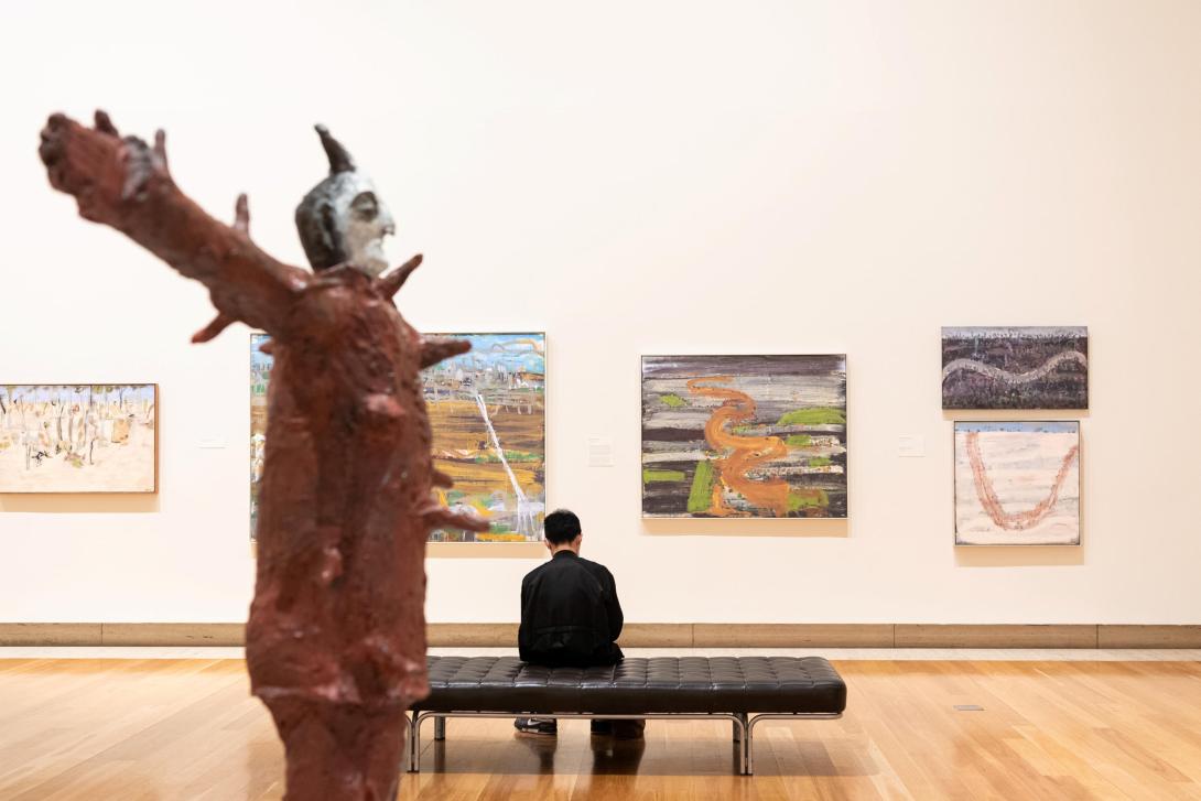 An installation view of several painted works hung on a white gallery wall, with a large bronze statue in the foreground. Between the two, a person sits on a bench facing the paintings.