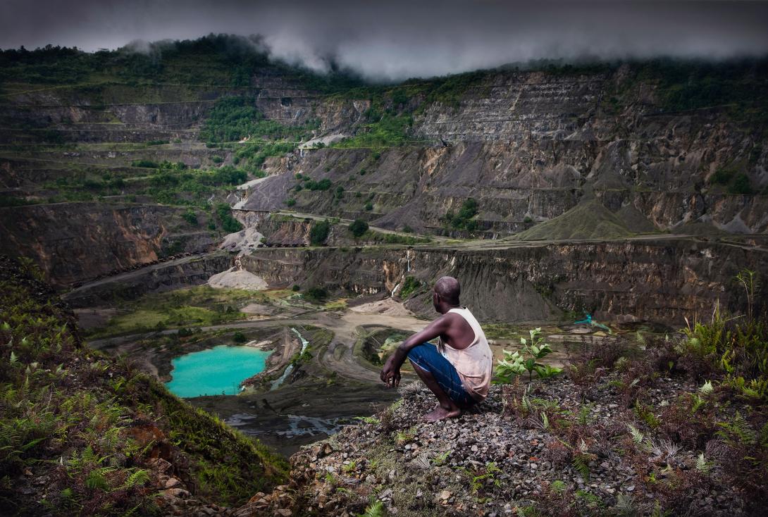 A photograph of a man squatting on a hill overlooking a quarry in a valley.