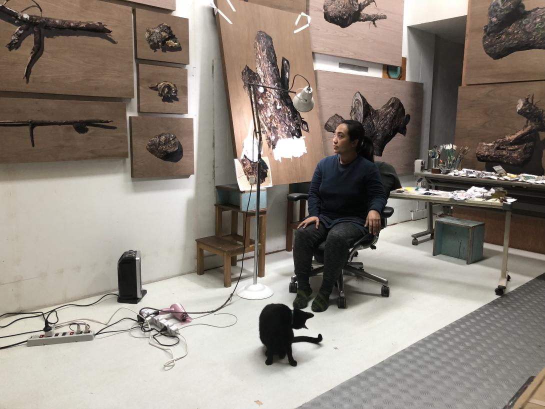 An artist sits on an office chair in her studio, looking at her portraits of pine trees. A black cat sits at her feet.