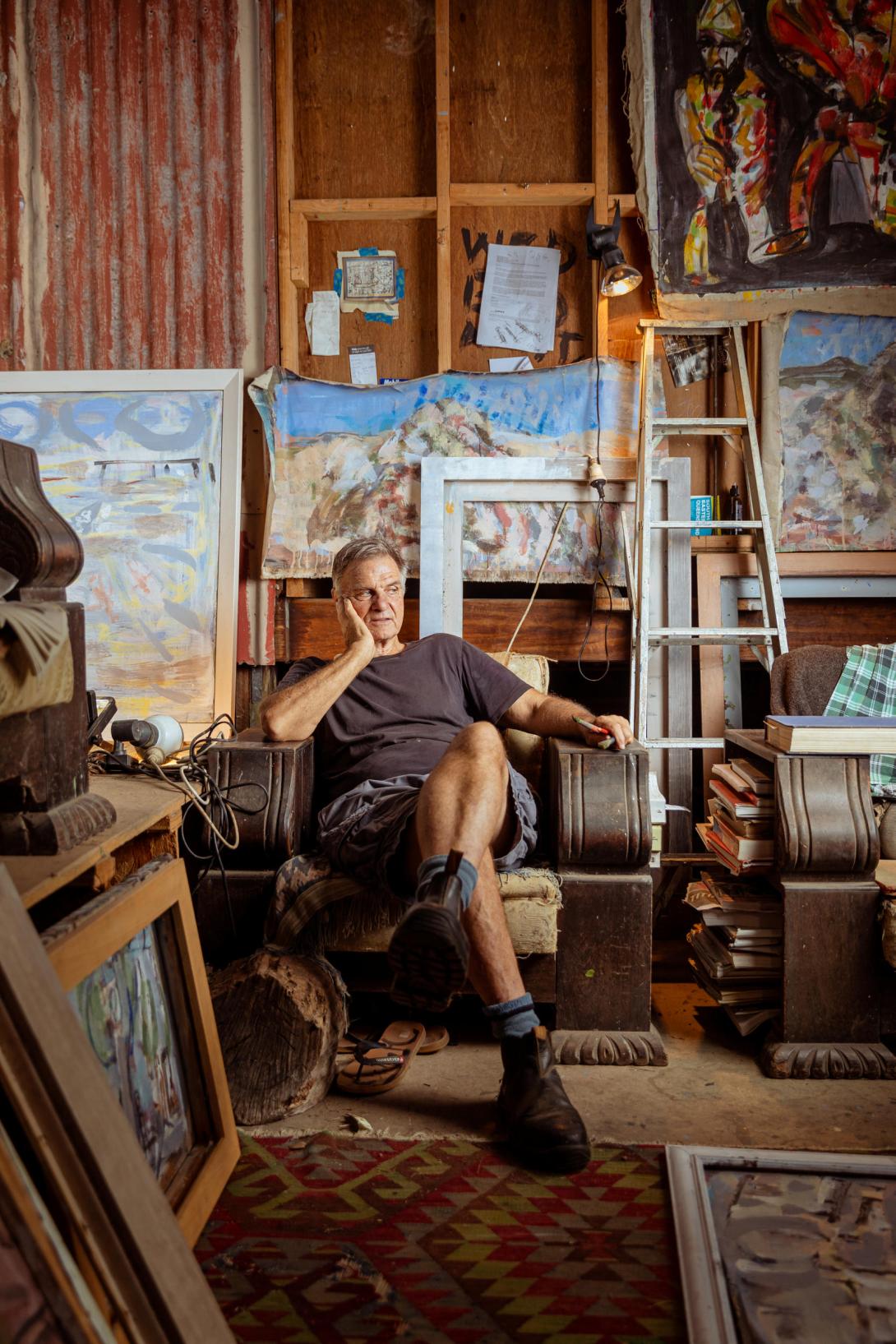A photograph depicting an artist; sits in a chair surveying his shed-studio, which is largely outside of the frame of the photograph. He is surrounded by works-in-progress, materials and tools.