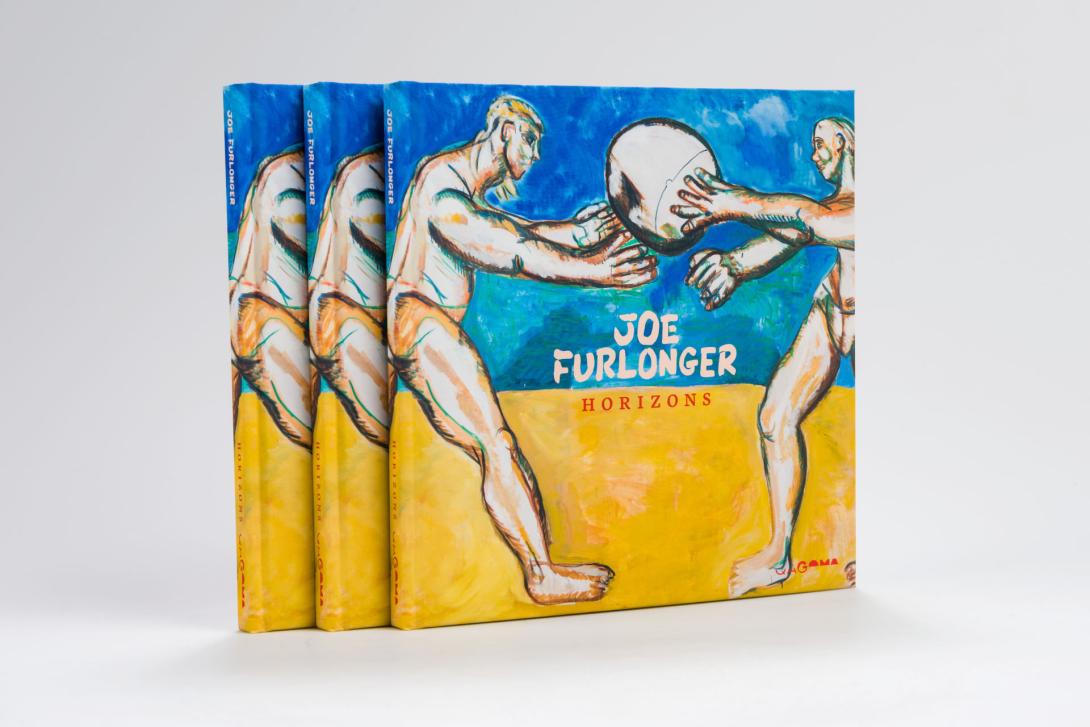A photograph of three copies of an exhibition catalogue for JOE FURLONGER: HORIZONS, featuring two volleyballers on a beach; the lower half of the cover depicts bright gold sand, and the upper half depicts a bright, deep blue sky.