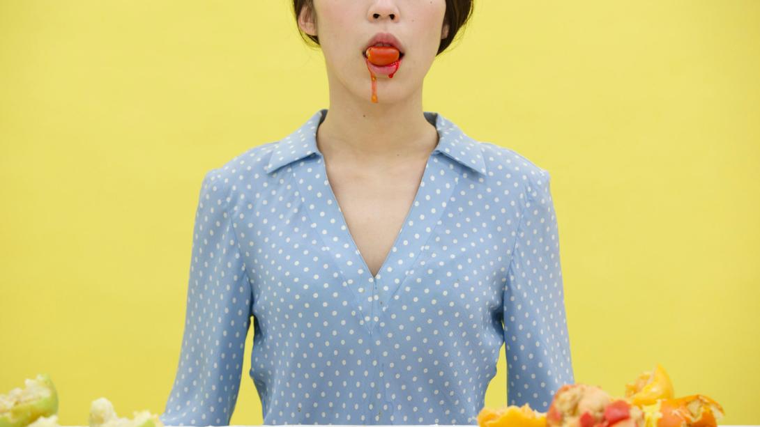 A photograph of a woman wearing a blue blouse and holding fruit against a yellow background. We only see from her nose to her elbows, and red fruit juice runs from her mouth.