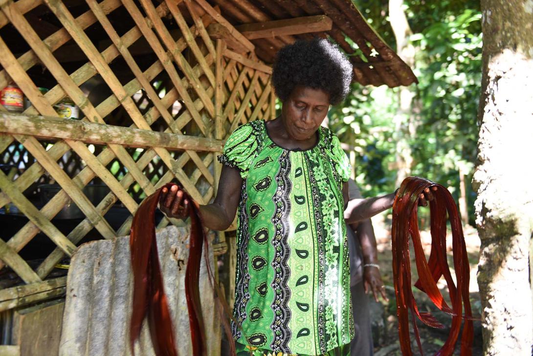 A woman wearing a green dress holds dark red-dyed pandanus thread in each hand.