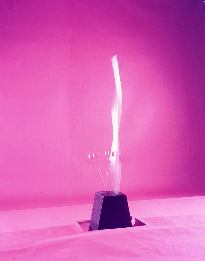 A sculptural work with kinetic elements stretching out of the top, which were moving when the photo was taken; their blurred movement is captured on a pink background.