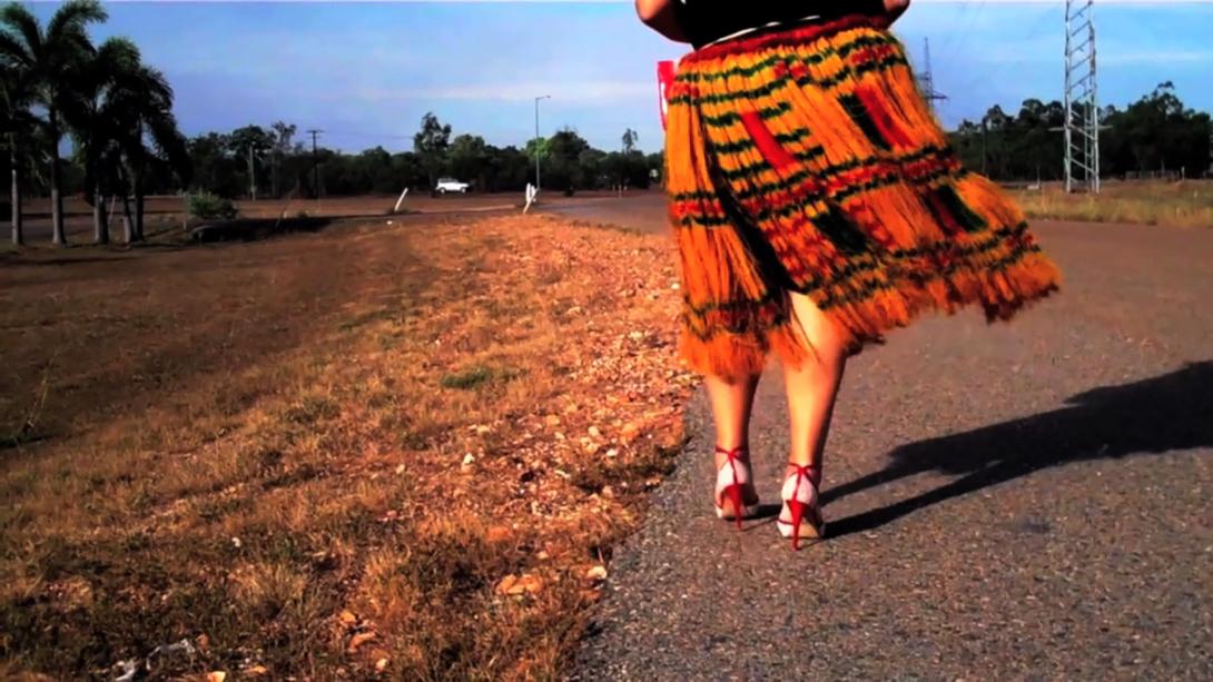 A still photograph of a woman in a red skirt and red high-heeled shoes walking down a country road, with orange dry grass at left.
