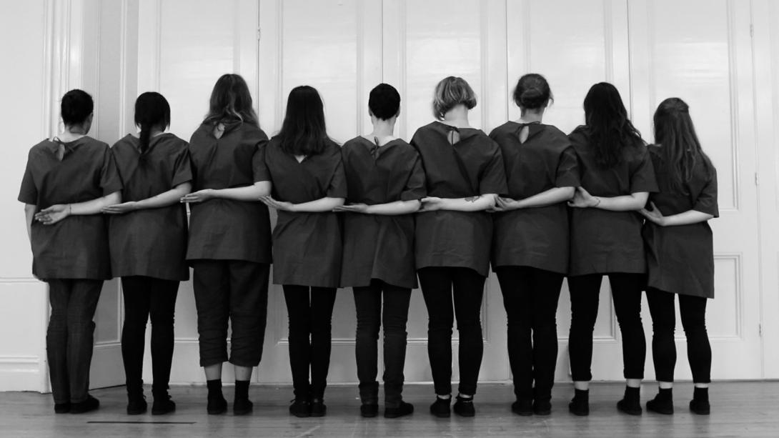 A still black-and-white photograph of nine women in matching shift tops standing in a row with their arms linked behind their backs, facing away from the camera.