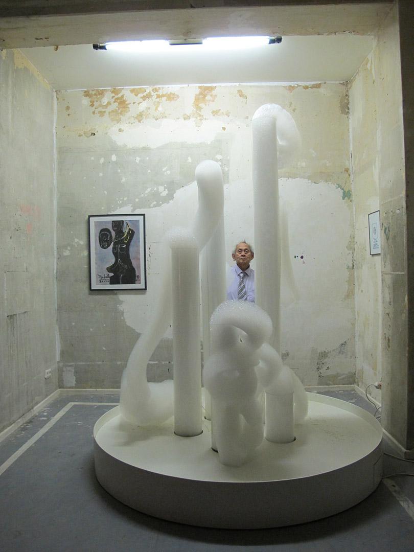 An installation view of Cloud Canyons, a work made of tubes and foam that bubbles and piles up organically, with the artist standing behind his work, so just his head and tie are visible.