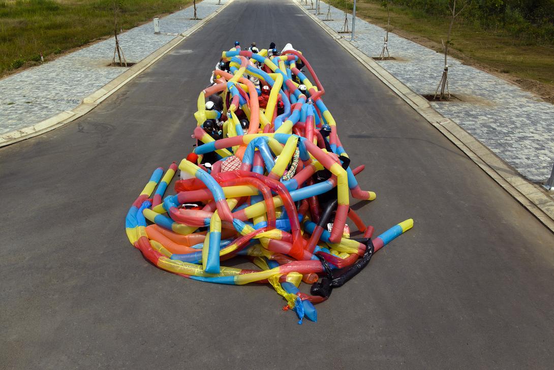 A photograph of a group of motorcycle riders with a web-like array of inflatable primary-coloured tubes stretched across them as they travel down a street.
