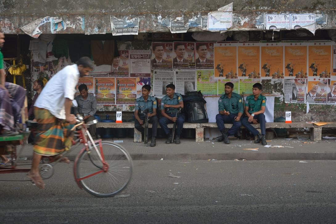 A photograph of a street scene in Myanmar, with someone riding a bicycle in the foreground and people sitting on a bench in the background.