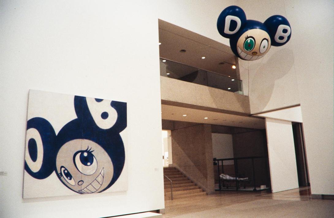 An installation view of two works installed in a gallery space at QAG. One is a painting of a cartoonish mouse-like face with large eyes and ears; another is a helium balloon version of this image, hung higher in the gallery.
