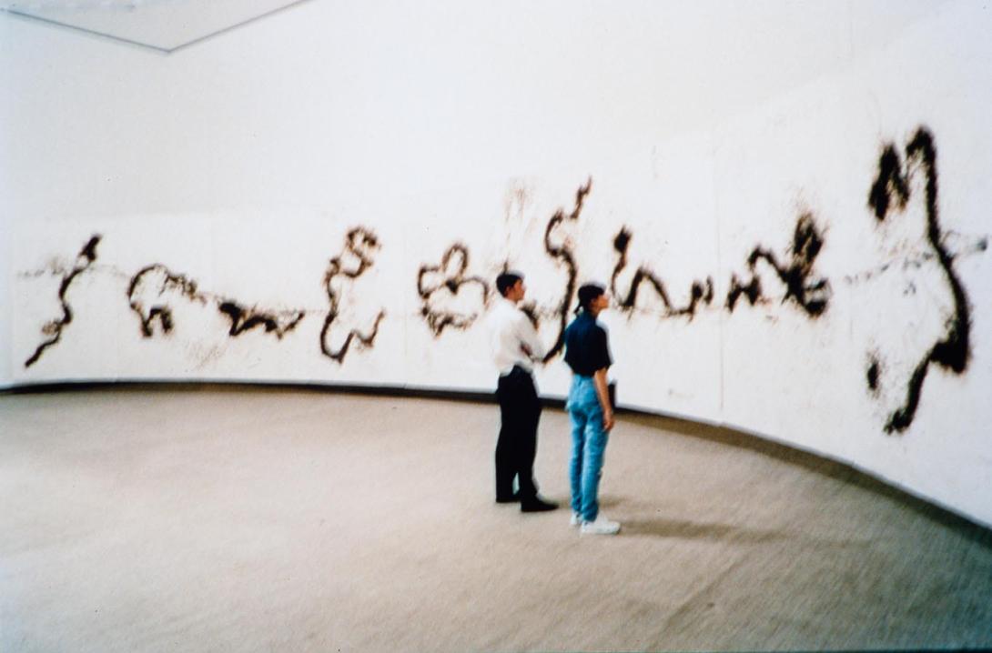 An installation view of a gallery space with a dragon- or river-link series of brown markings over a curved wall. Two visitors stand in the gallery, looking at this work.