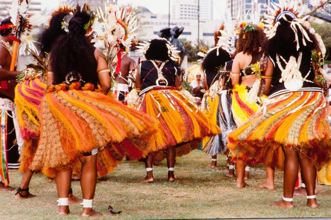 A photograph of a group of Papua New Guinean dancers wearing orange skirts that sway and twirl; the photograph is taken with the dancers facing away from the camera, facing the Brisbane River.