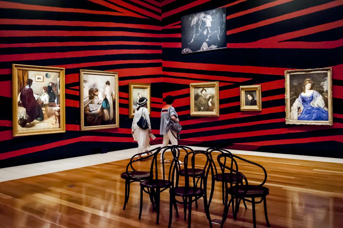 An installation view of works in gold frames from QAGOMA's Australian Collection installed on a red-and-black striped background in a gallery space with polished hardwood floors.