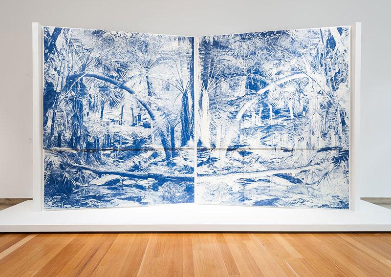 An installation view of two panels depicting a forest in blue pastel and pencil.
