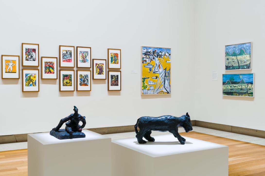 An installation view of small framed works alongside larger paintings in a gallery space, with two small bronze sculptures on plinths in the foreground.