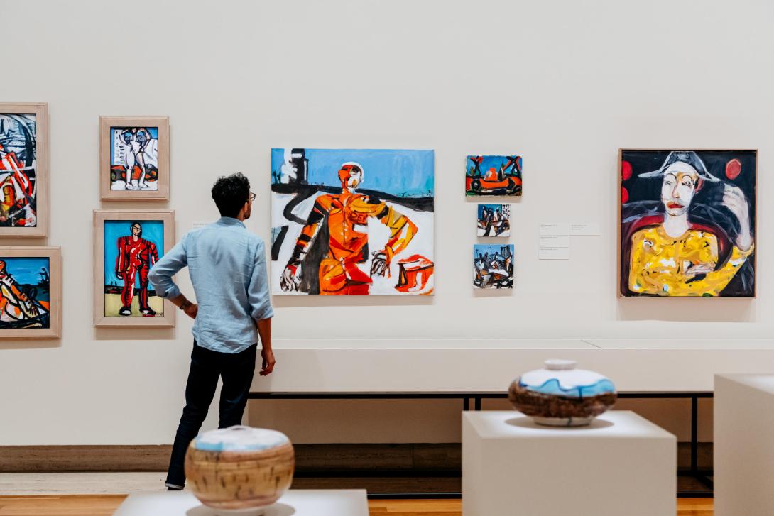 An view of 10 works installed in a gallery space: eight are paintings of various sizes hung on the white wall, while two are ceramic pots on plinths. A man in a blue shirt faces the paintings, looking closely at one in the centre.