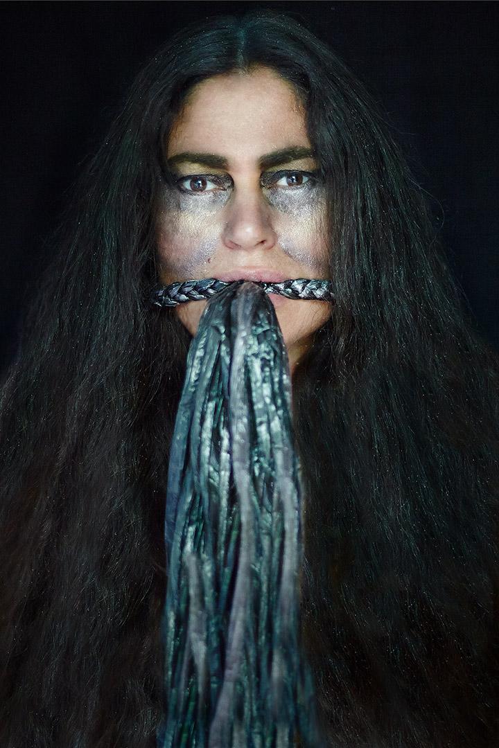 A photograph of a woman with long, dark hair; the background of the photo is completely black, and the woman has glitter make-up on and under her eyes. A shiny material is braided like a bit at the woman's mouth, with tassels of the shiny material appearing to flow from her mouth.