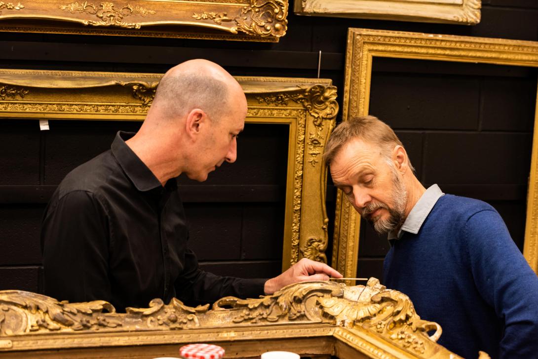 Two men inspect the corner of an ornate gold frame in the QAG framing workshop.
