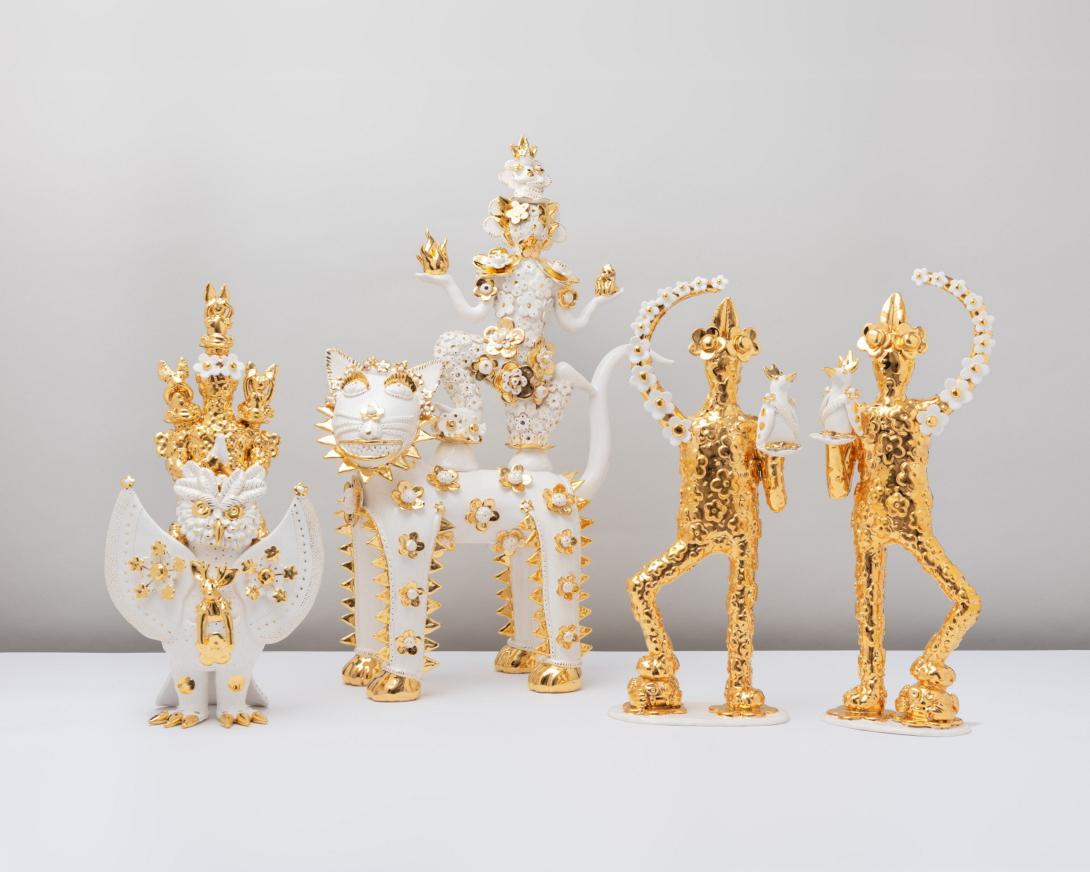 Four gold-and-white ceramic figures against a white background: an owl, a person standing atop a cat, and two figures with birds on their shoulders.