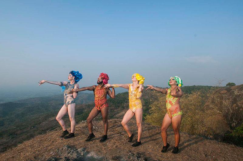 In this photograph, four drag queens stand on a high hill overlooking a valley; they wear blue, red, yellow and green respectively.