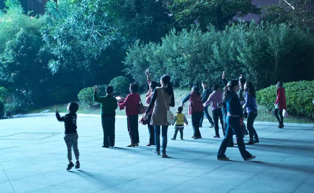 Standing in evening light on pavement, a group of people facing away from the camera wave at someone or something out of frame.