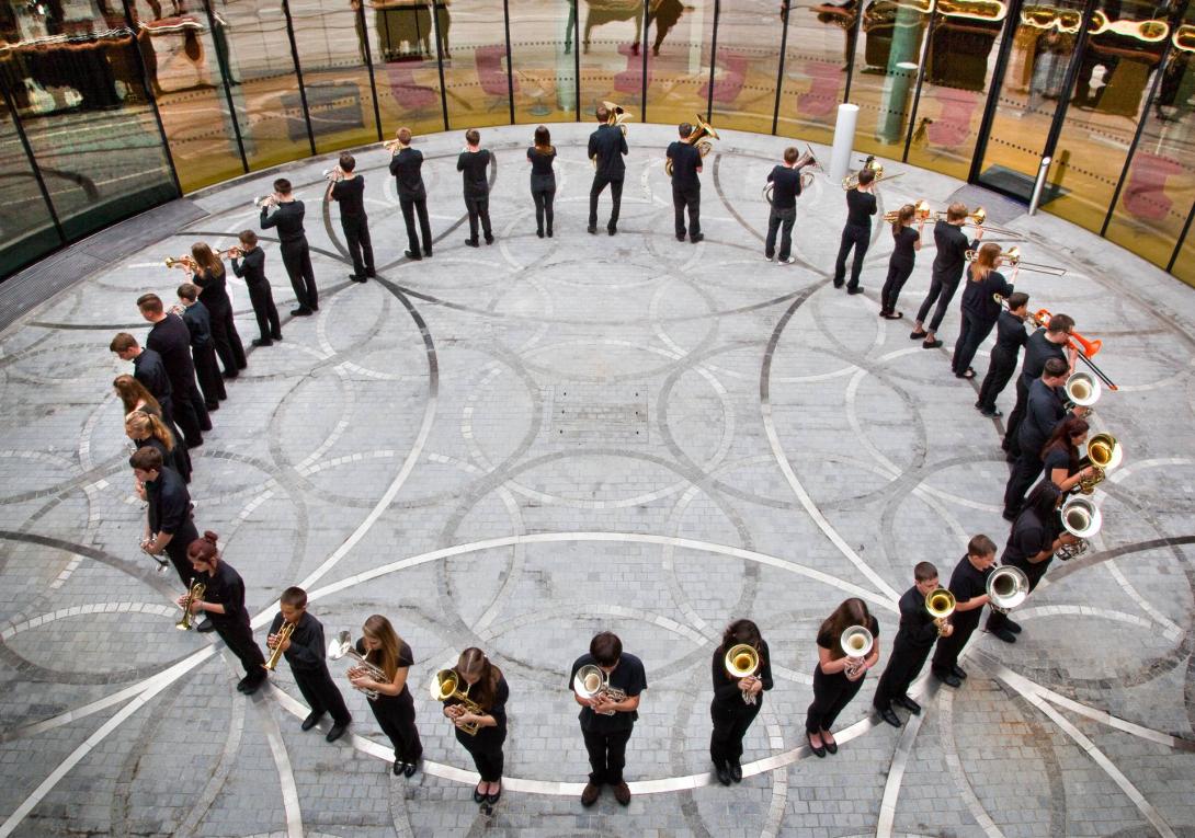 A photo of a brass band, all dressed in black, standing in a circle; facing outwards, standing on decorative pavement, they play their instruments.